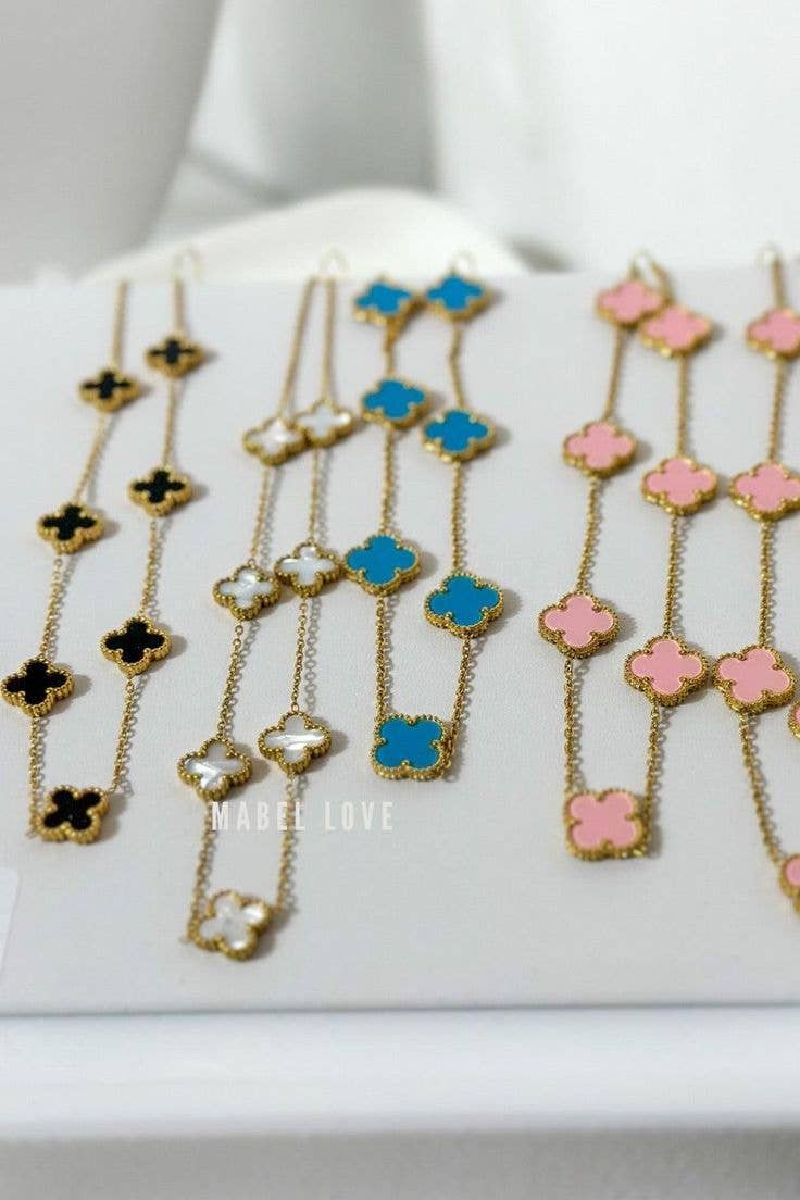 Three gold-tone chain necklaces with clover-shaped charms in black, white, blue, and pink, displayed on a white jewelry stand.