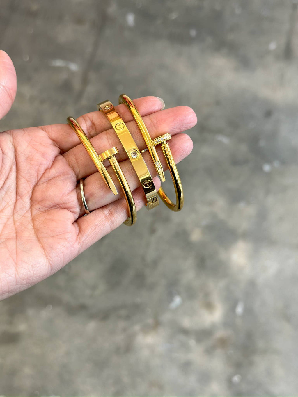A hand holding several slim gold bangle bracelets against a blurred gray concrete background.